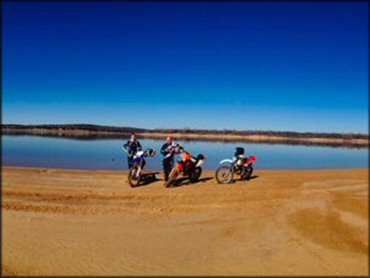 Two men standing next to a KTM and two Honda dirt bikes parked next to lake.