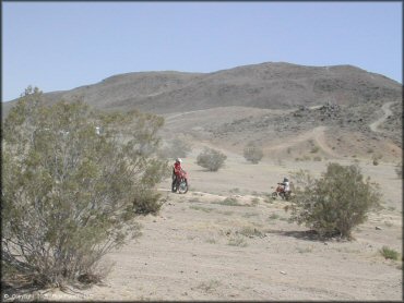 Two Honda dirt bikes riding around flat area.