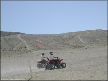 Two Honda ATVs each with red whip flags and one carrying a cooler strapped to the rear.