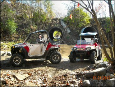 Two Polaris RZR 800 UTVs with orange whip flags parked next to shallow stream.