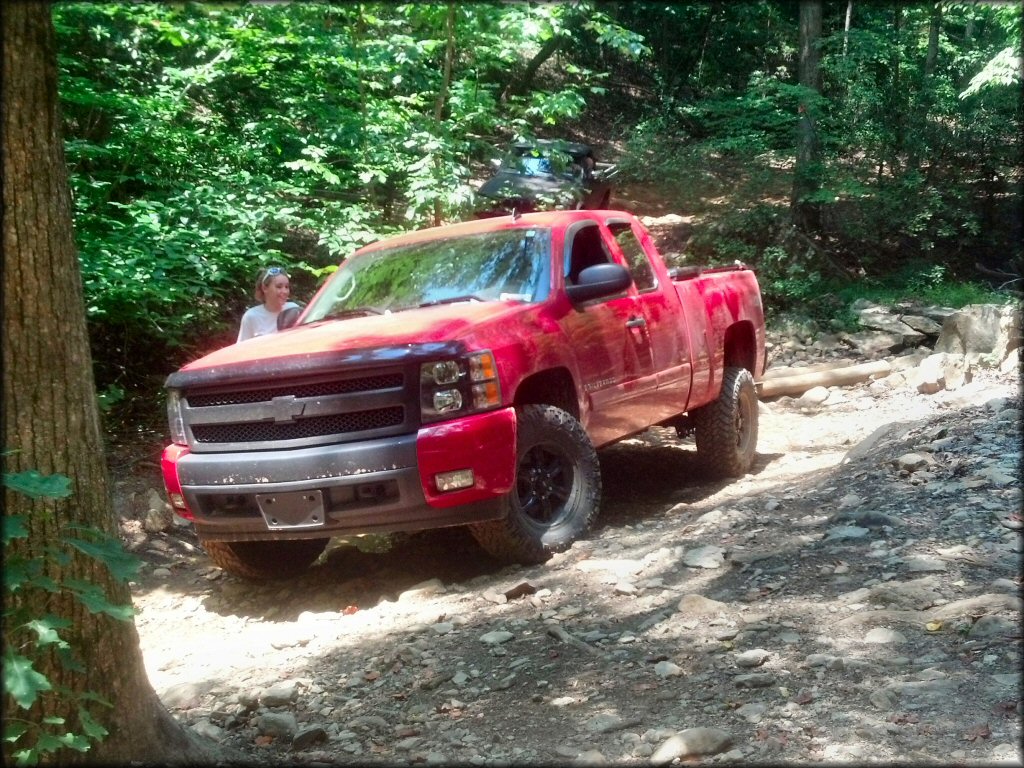 Red Chevy pickup truck with off-road tires and black alloy rims on a rocky dirt trail with a woman nearby.