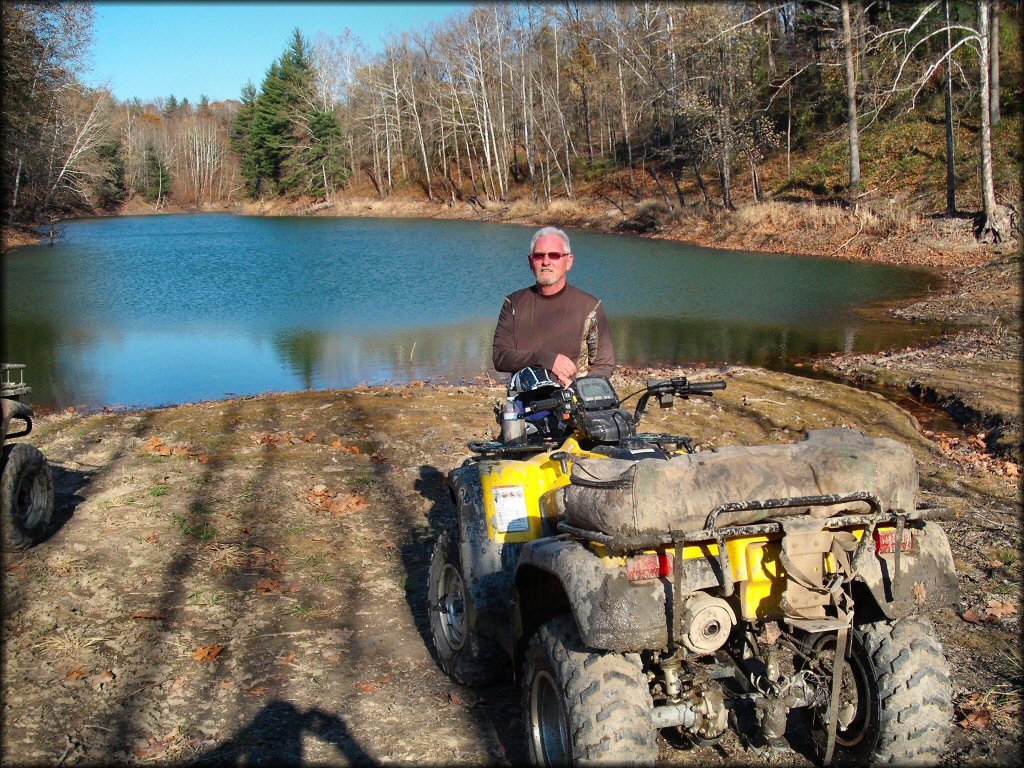 Mature man standing in front of mud covered Suzuki ATV with scenic lake in the background.