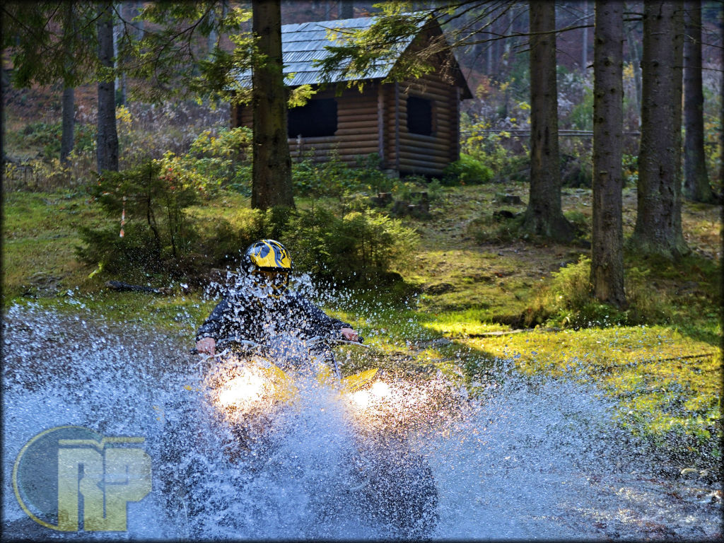 A Can-Am ATV crossing a stream in the woods with a cabin in the background