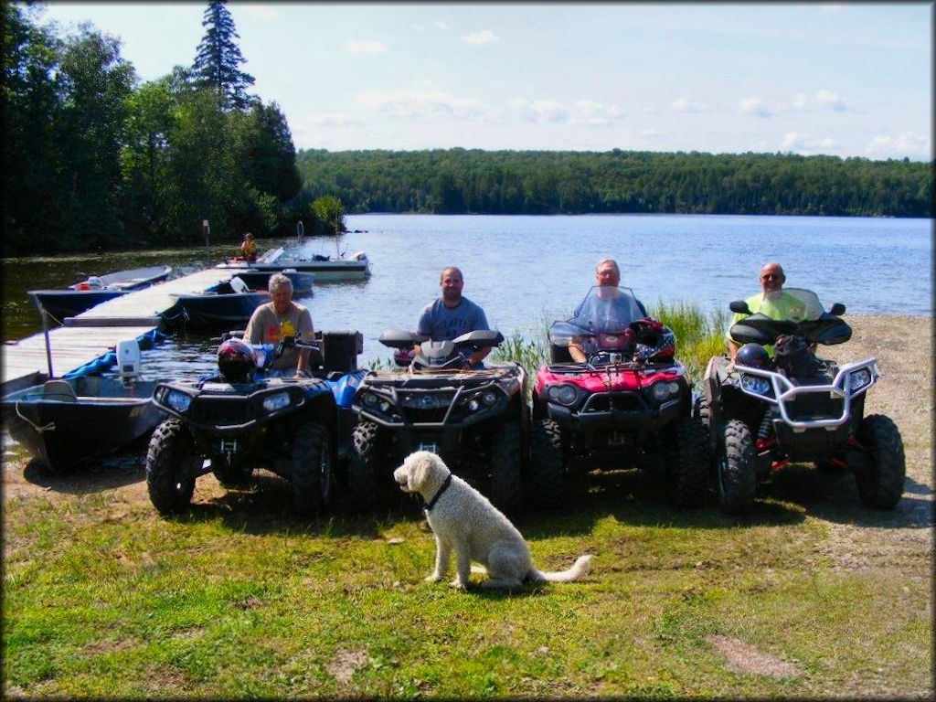 Group of mature men sitting on ATVs next to dock with boats in the background.
