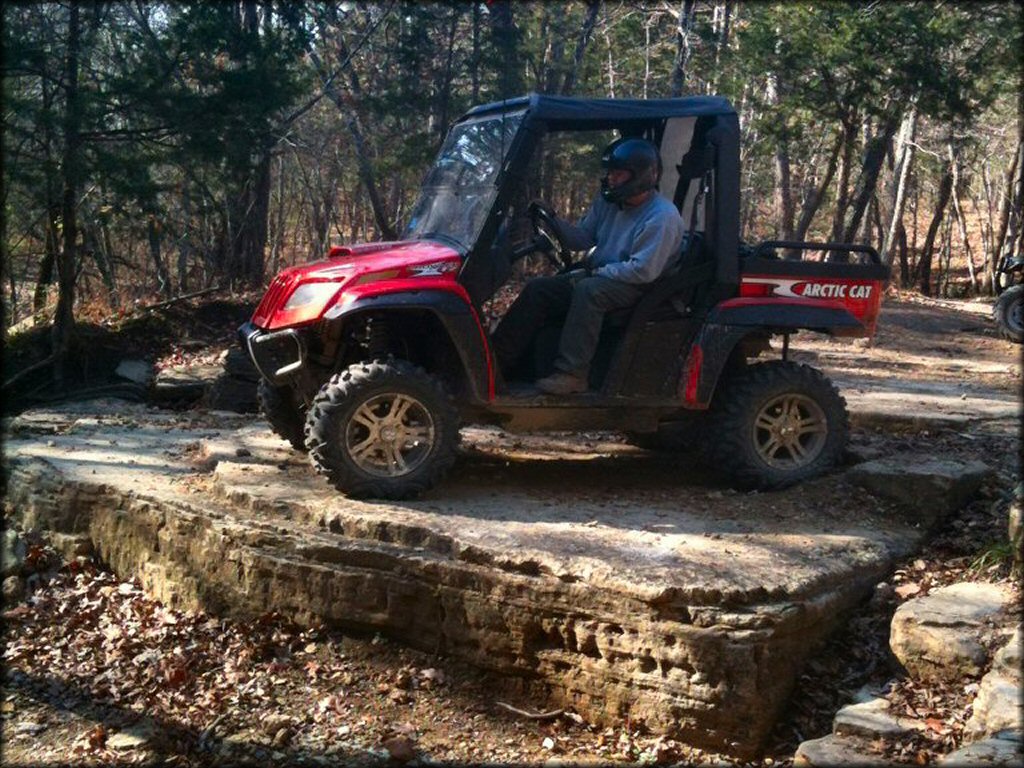 Young man sitting in drivers seat in Red Artic Cat with black UTV roof parked alongside rocky trail.