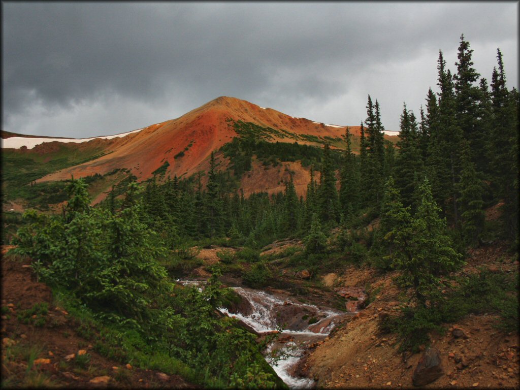 Scecnic photo of reddish colored mountain with pine trees and shallow creek at the base.