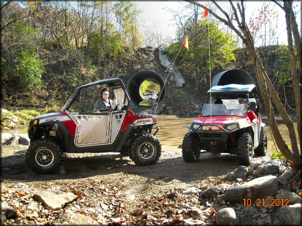 Two red Polaris RZR 800 UTVs with orange whip flags parked next to shallow creek with drainage tunnels in background.