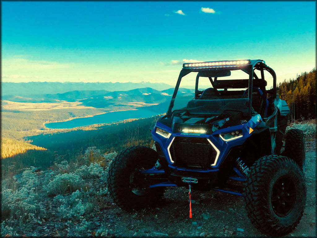 Blue Polaris UTV with LED Light Bar parked alongside trail with lake and mountain views in the background.