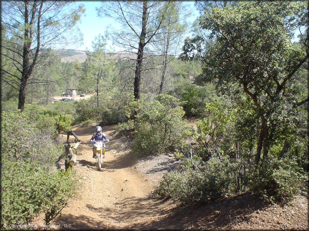 Rider on Suzuki RM100 going up trail with campground in the background.