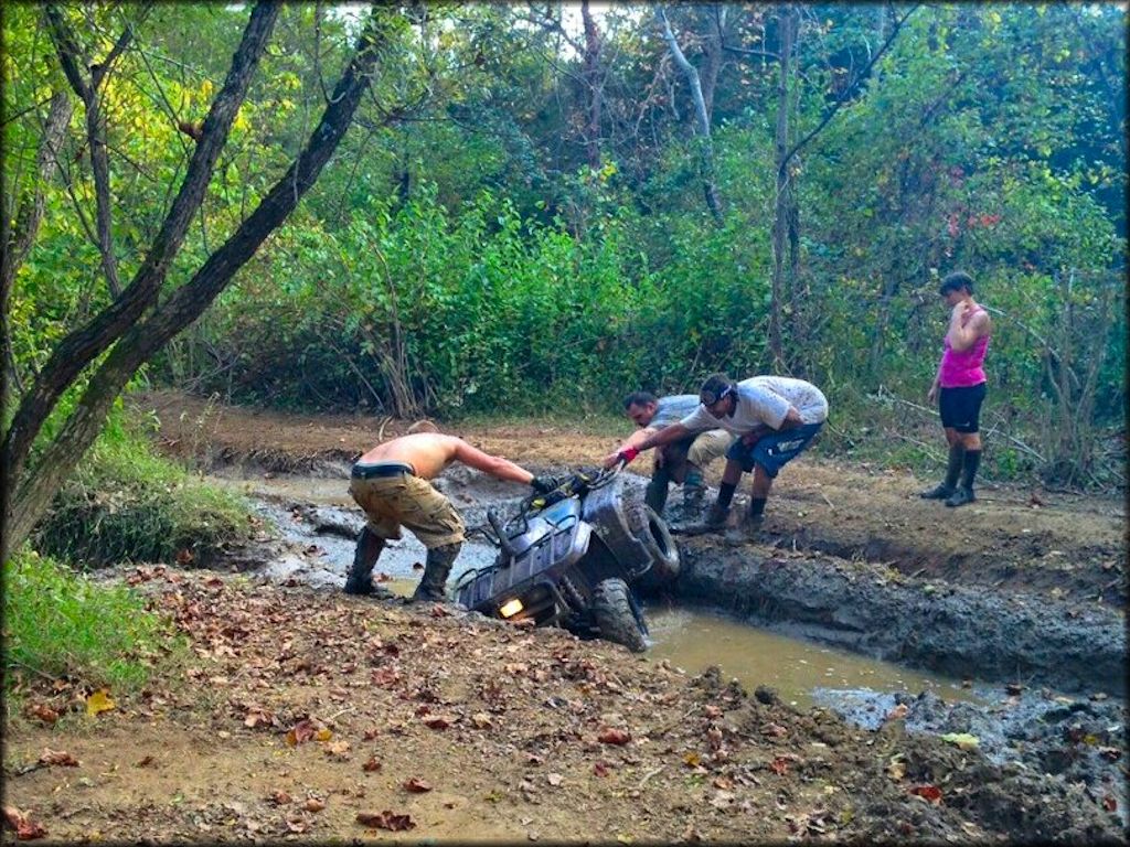 Three men trying to pull ATV from deep muddy rut.