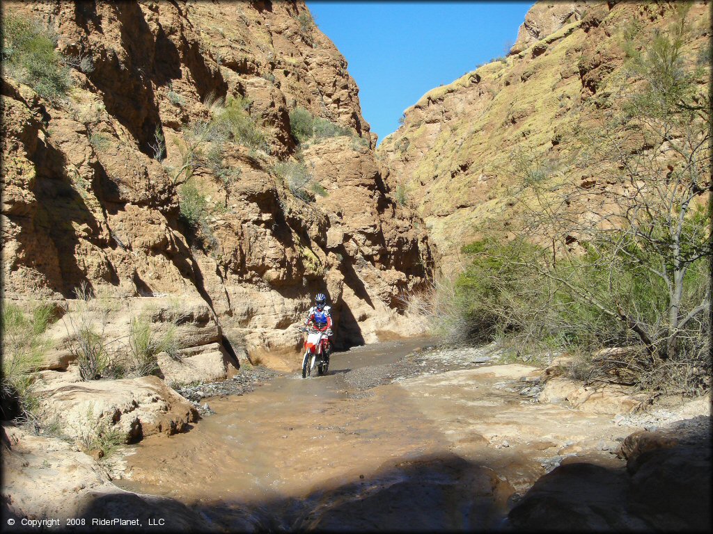 Honda dirt bike going through stream with gravel wash and flat river rocks.