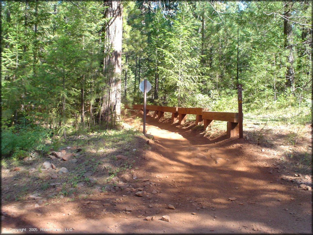 Stop sign with wooden guard rail at intersection of trail and paved road.