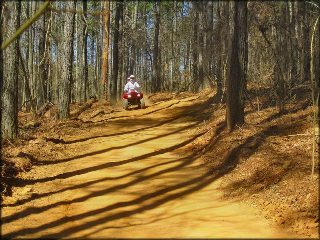 Two people riding a Honda four wheeler down wide and hard packed offroad trail.