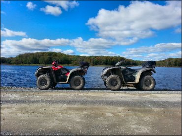 One Red KYMCO 4x4 500 and one Green KYMCO 4x4 ATVs with rear saddle bags parked next to lake.