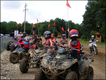 Large group of riders sitting on ATVs with orange whip flags parked in staging area.