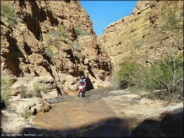 Honda dirt bike going through stream with gravel wash and flat river rocks.