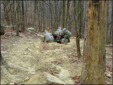 Two people pushing ATV thorugh rocky section of trail.