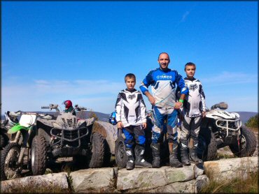 Man and two young boys standing next to three Yamaha ATVs and one Kawasaki dirt bike.
