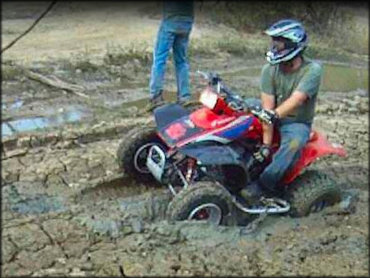 Young man wearing helmet and riding gloves sitting on ATV that is stuck in the mud.