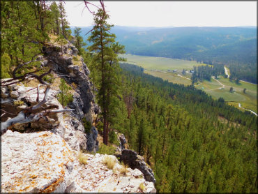A close up photo of steep cliff overlooking pine tree forest and valleys.