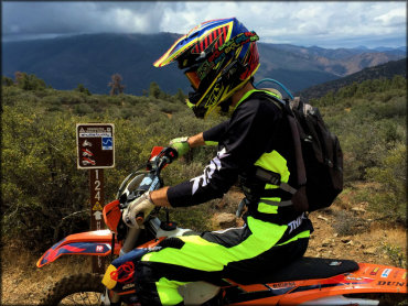 A close up photo of a KTM dirt bike and rider parked next to forest service carsonite trail marker.