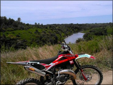 Husqvarna dirt bike with FMF exhaust pipe parked off the trail with grassy hills and small lake in the background.