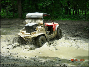 Red Polaris side by side with black solid roof carrying ice chest and tool box going through mud puddle.