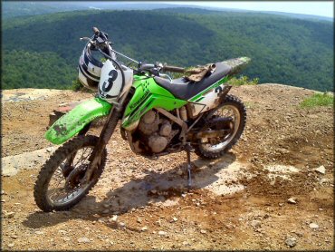 Kawasaki four-stroke dirt bike parked with helmet on handlebars and gloves on seat overlooking scenic view.