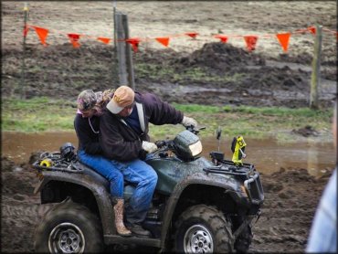 Man and woman riding on a dark green Honda ATV going through muddy section.
