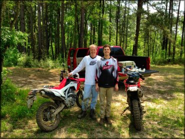 Father and son with two Honda dirt bikes and red pickup truck parked at trailhead.