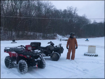 Man wearing cold weather coveralls and motorcycle helmet standing next to two Polaris Sportsman ATVs in the snow.