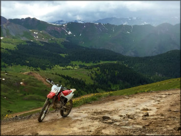 A Honda CRF 230 parked on a ATV trail with green meadows and snow capped mountains in the background.