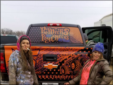 Two young women covered in mud standing behind rear of Chevy truck.