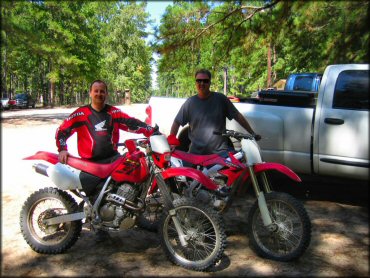 Two men standing behind a Honda XR, Honda CRF and other trucks parked in staging area.