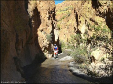 Scenic view of Honda dirt bike riding through small runoff stream flowing through colorful box canyon.