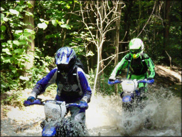 Two dirt bike riders on blue Yamaha motorcycles wearing Moose and Fox trail riding gear going through water crossing.