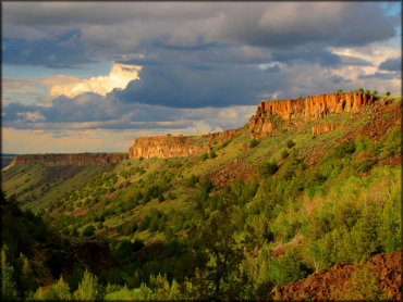 A scenic photo of red orange rock mesas surrounded by green meadows and various trees taken from the trail.