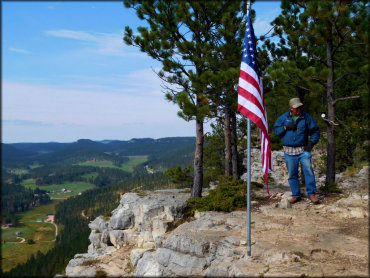 Elderly man standing next to flagpole with American flag overlooking cliff with scenic view of valleys, meadows and forests.