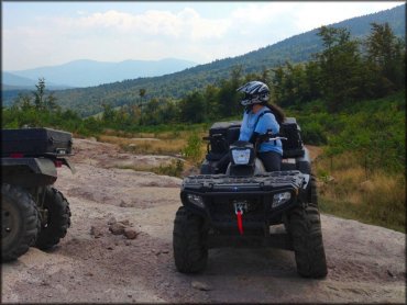 Woman sitting on Arctic Cat ATV with mud tires, rear cargo storage boxes and winch with red tow strap attached to front bumper.