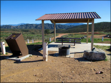Photo of shade gazebo on concrete pad with picnic table, fire ring and trash receptacle.