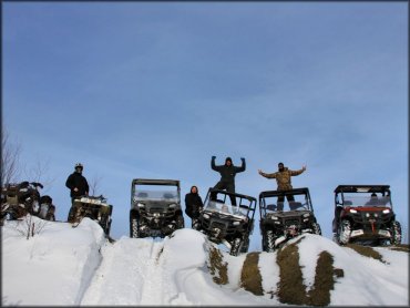 Four men wearing cold weather riding gear standing next to four UTVs and two ATVs parked on snowy hill.