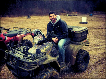 Man sitting on top of parked Polaris 700 Sportsman ATV with mud tires and rear storage box.