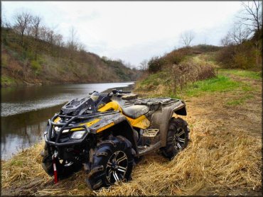 Can-Am Outlander X with winch and red tow strap on front bumper and mud tires parked on grassy bank next to lake.