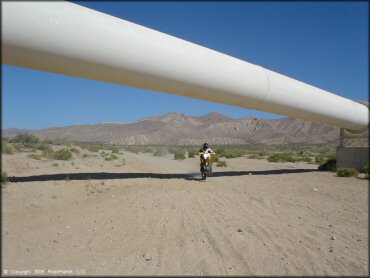 Man on Suzuki RM-250 popping a wheelie while riding underneath one of the Los Angeles Aqueduct pipelines.