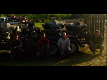 Group photo of four men kneeling in front of mud covered ATVs on flat bed trailer.