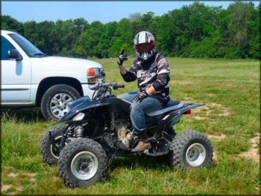 Rider wearing helmet and motocross jersey sitting on Yamaha Raptor parked next to white truck in grassy meadow.