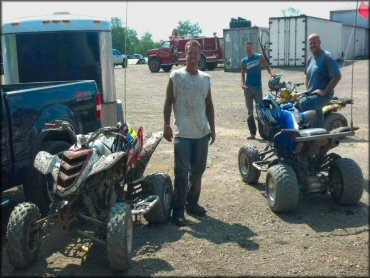 Three men standing next to two Yamaha Raptor ATVs and one Suzuki ATV with whip flags parked next to box trailer.