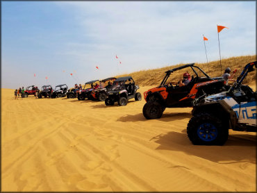 Large group of UTV with hard top roofs and orange whip flags parked in the sand.