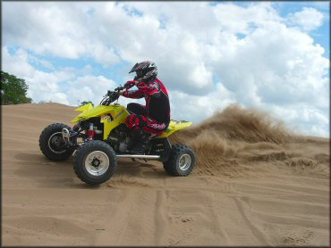 Man wearing black and red Fly Racing motocross gear riding Suzuki ATV through sand dunes.