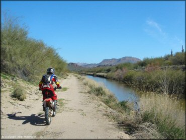 Woman wearing Fox Racing motocross gear and CamelBak riding Honda CRF-150 alongside canal.